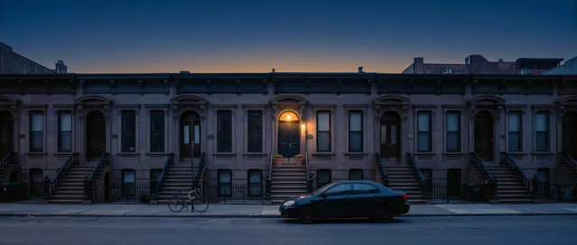 A row of matching brownstone townhouses at blue hour with one porch light on above one front door